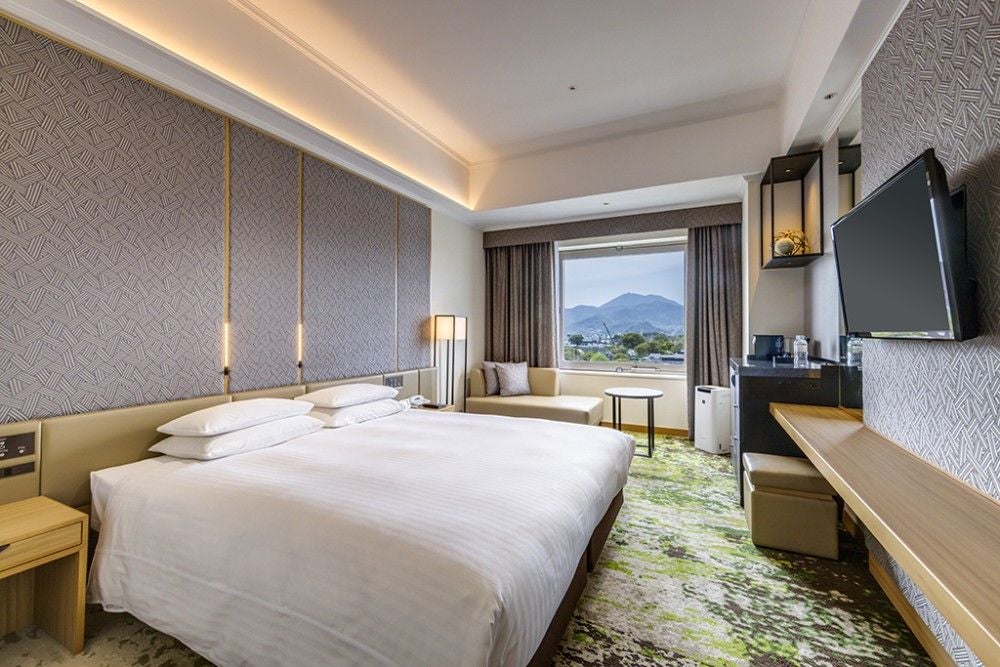 Modern king room with city views, featuring plush white bedding, dark wood furnishings, and floor-to-ceiling windows in Hotel Nikko Kumamoto