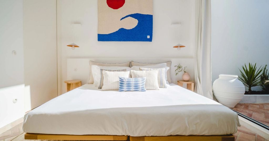 Bright, minimalist hotel room with whitewashed walls, arched window, woven pendant lamp, and rustic wooden furnishings overlooking Lagos town