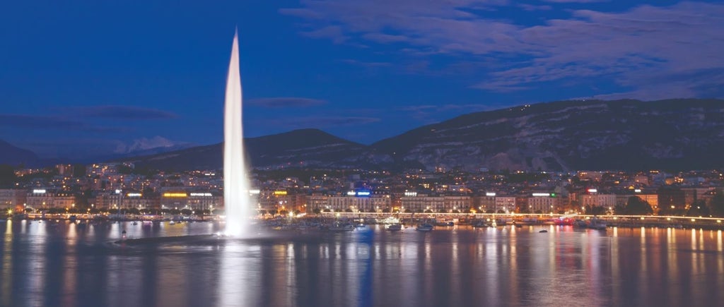 Majestic Fairmont Grand Hotel Geneva overlooking Lake Geneva, with elegant glass facade and ornate architecture reflecting in calm waters