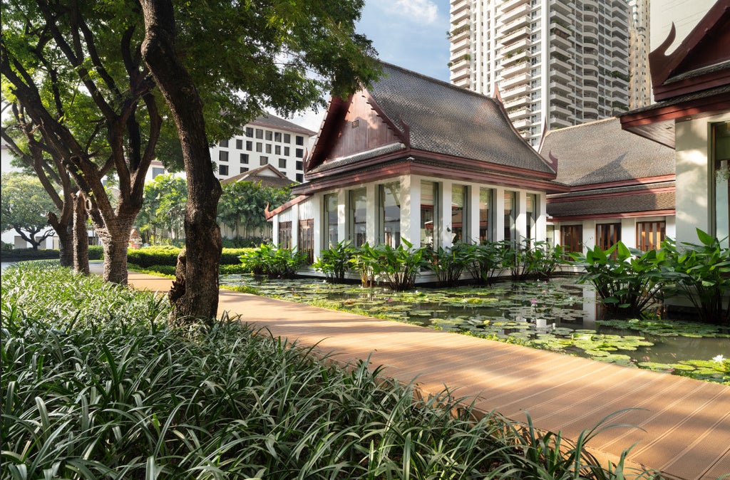 Elegant Thai-style hotel exterior with lotus ponds, stone pathways, and lush tropical gardens surrounded by modern skyscrapers at dusk