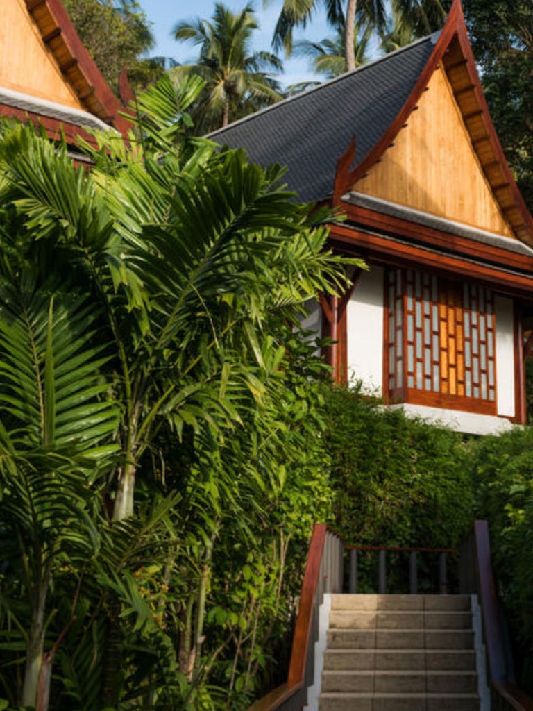 Elegant Thai pavilion room with dark wood furnishings, white linens, floor-to-ceiling windows overlooking lush tropical gardens