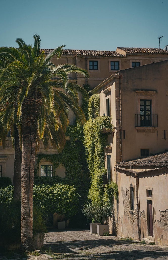 Elegant historic Sicilian hotel with stone facade, balconies overlooking narrow street, warm golden sunlight illuminating traditional Mediterranean architecture