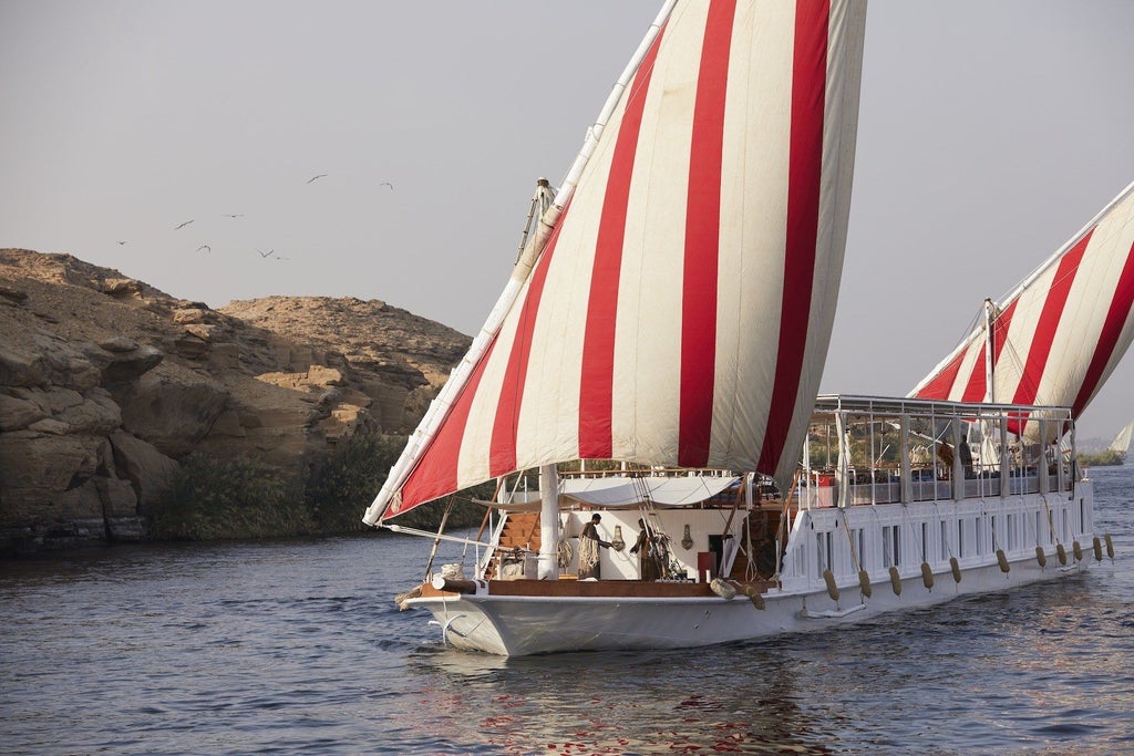 Traditional wooden dahabiya boat with cream-colored sails moored on the Nile, featuring elegant canopied deck and ornate Arabic-style details.