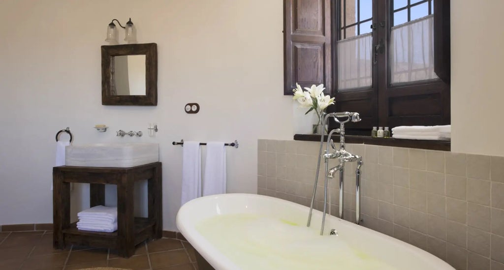 Elegant rustic suite in white-washed stone walls, featuring wooden beamed ceiling, antique furnishings, and soft natural light through arched windows