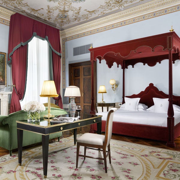 Ornate Italian hotel suite with crystal chandelier, gilded gold mirrors, antique furnishings, and plush red drapes overlooking gardens