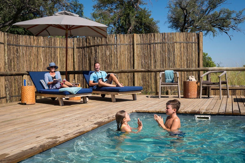 Elevated safari tent lodge with wooden deck overlooking flooded Okavango Delta, surrounded by palm trees at golden hour