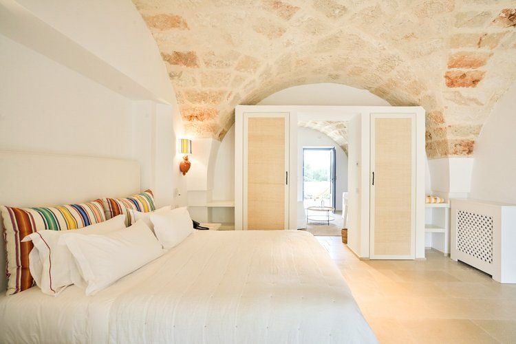 Elegant stone-walled suite with rustic wooden furniture, crisp white linens, and panoramic windows overlooking Puglia's serene countryside landscape