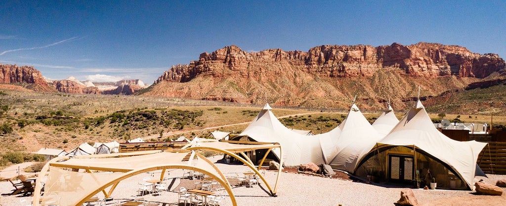 Luxury safari-style tent nestled against rust-colored rock formations of Zion National Park, with golden sunset light illuminating canvas and landscape