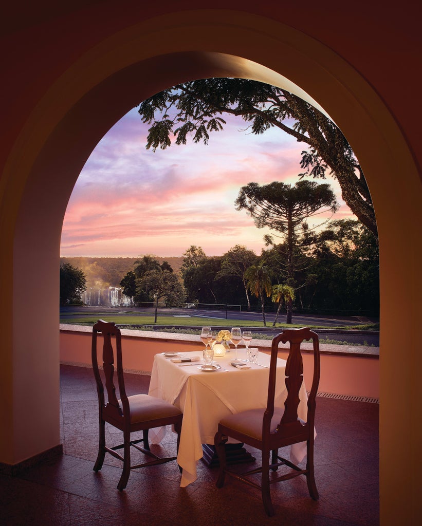 Luxurious pink-hued colonial hotel building with elegant archways nestled beside Iguazu Falls, surrounded by lush tropical gardens