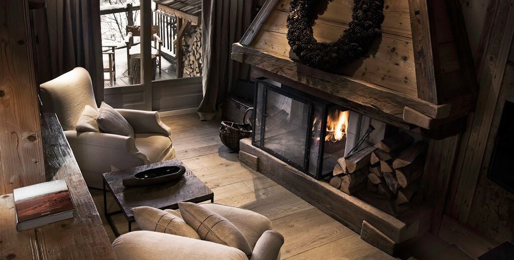 Elegant alpine suite with warm wooden interiors, plush white bedding, and floor-to-ceiling windows overlooking snow-capped mountain landscape in French luxury chalet