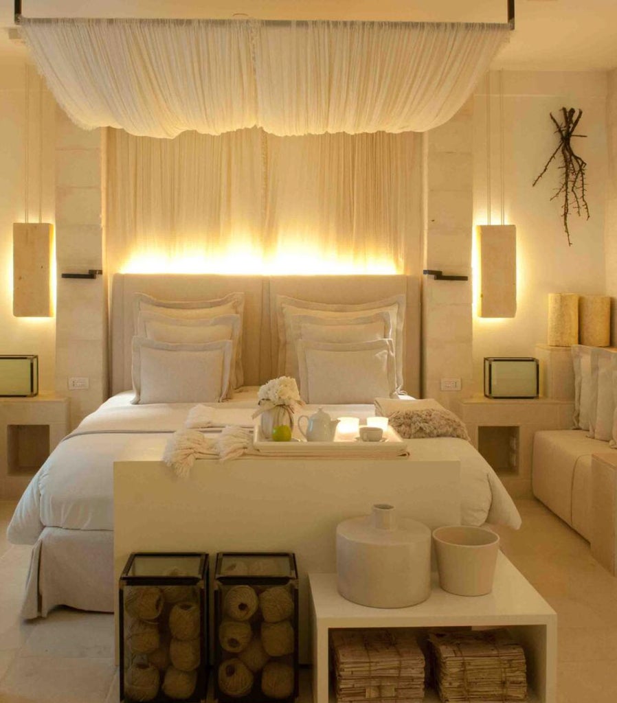 Elegant Mediterranean hotel room with stone walls, arched windows, white linens, woven rug and rustic wooden furniture bathed in natural light