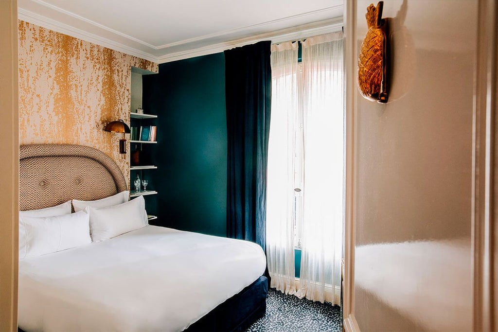 Elegant Parisian hotel room with plush king bed, rich navy walls, vintage brass accents, and large window overlooking charming street in boutique Grand Pigalle Hotel