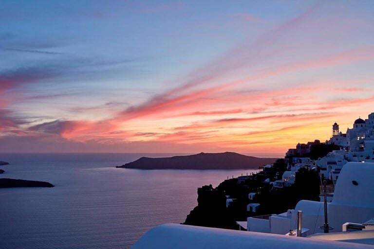 Luxurious white-washed suites of Chromata hotel cascading down Santorini's iconic cliffside with stunning azure Aegean Sea panorama at sunset