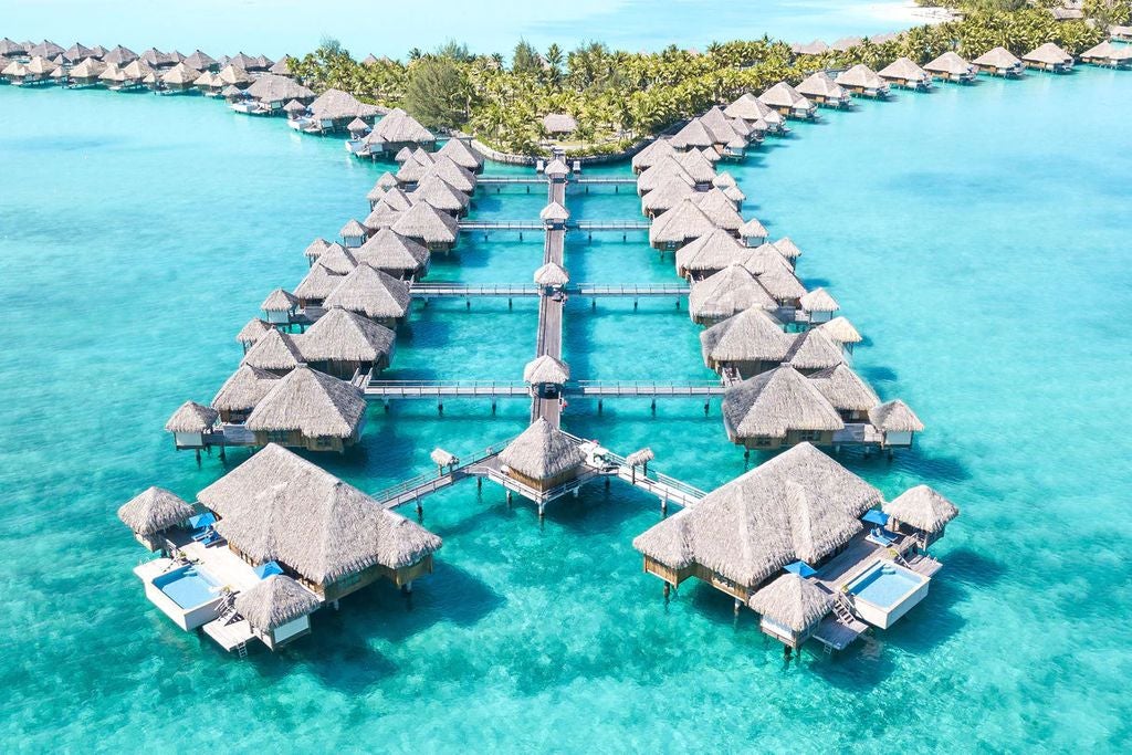 Luxurious overwater villas with thatched roofs stretching across crystal-clear turquoise lagoon, backed by lush mountains in Bora Bora.