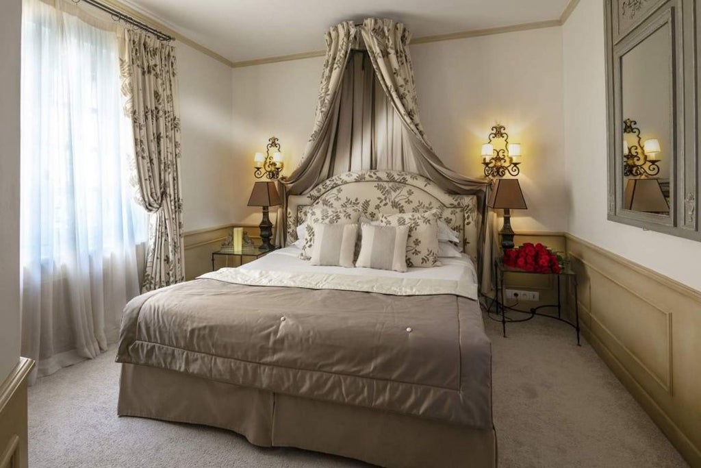 Elegant deluxe hotel room with plush white bedding, soft gray walls, modern wooden furnishings, and large windows overlooking charming French countryside scenery