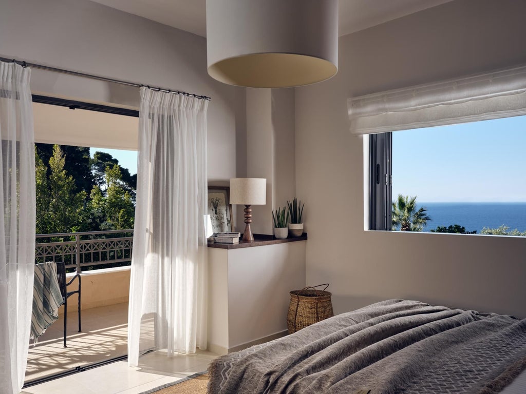 Spacious Mediterranean-style villa bedroom with elegant white furnishings, wooden floors, and panoramic sea view through large arched windows overlooking azure coastline.