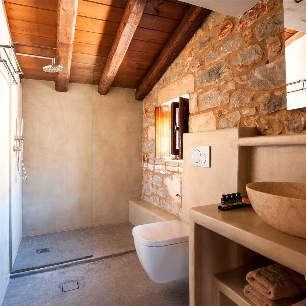 Elegant loft suite with stone walls, wooden beam ceiling, white linens, and panoramic coastal view of scenic Greek landscape through large windows