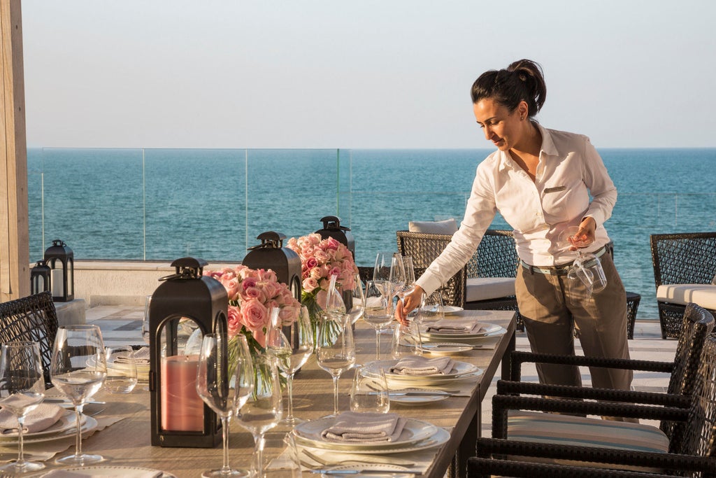 Elegant Mediterranean-style luxury hotel with white arches and domes overlooking turquoise sea, palm-lined courtyard at sunset