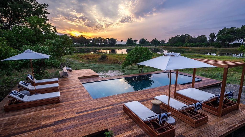 Elegant safari lodge with thatched roof and wooden deck overlooking a serene Okavango Delta lagoon at golden hour