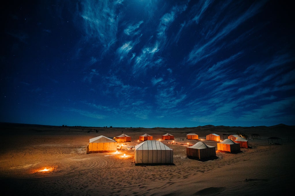 Elegant Moroccan desert camp tent with ornate carpets, lanterns and plush furnishings nestled among rolling sand dunes at sunset