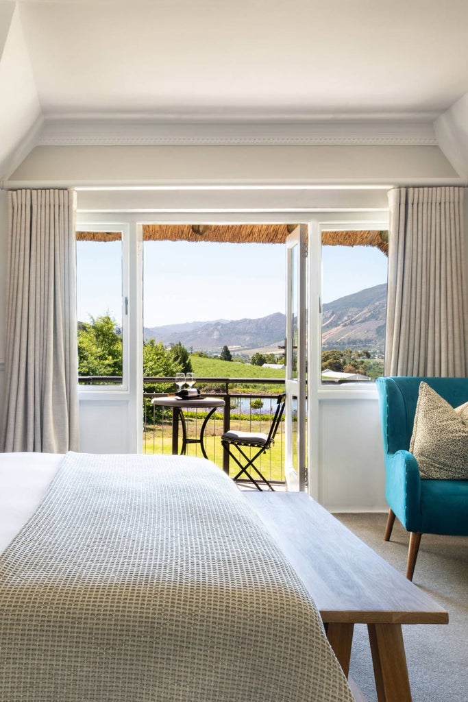 Elegant king-size bed with plush white linens, dark wood furnishings, and French doors overlooking vineyards at luxurious Mont Rochelle hotel room in South African wine country