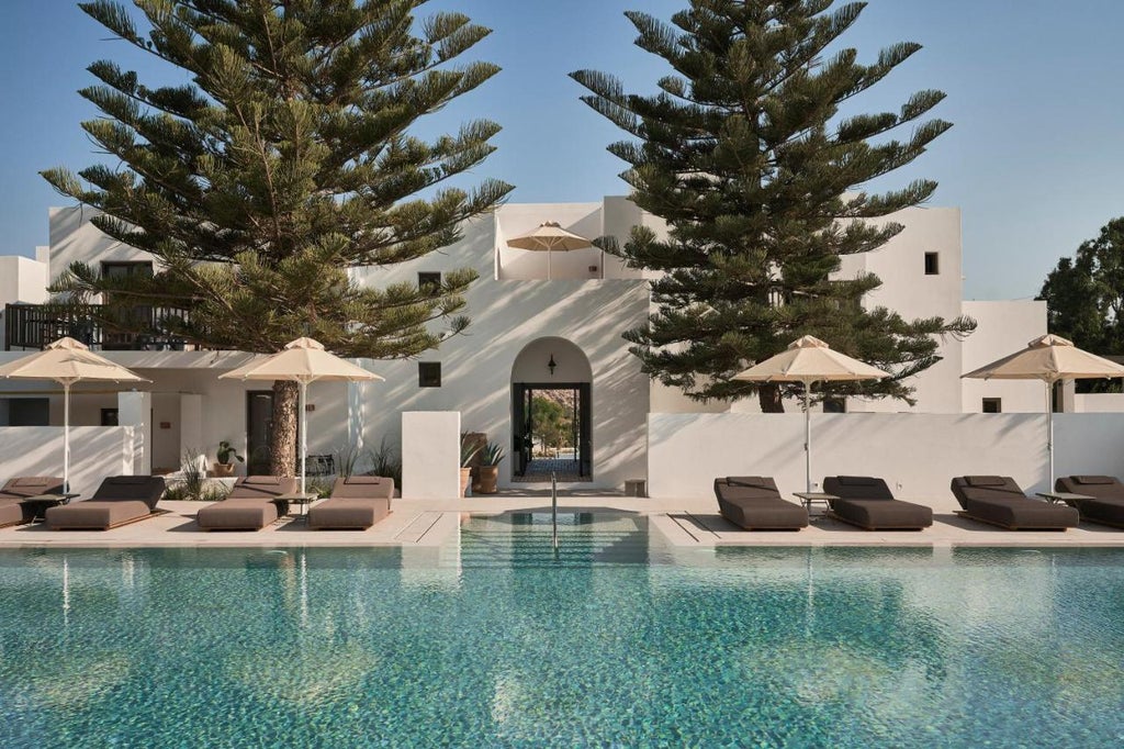 White-washed Cycladic luxury hotel building with curved archways, set against rocky hillside and blue Greek sky, stone paths meander through