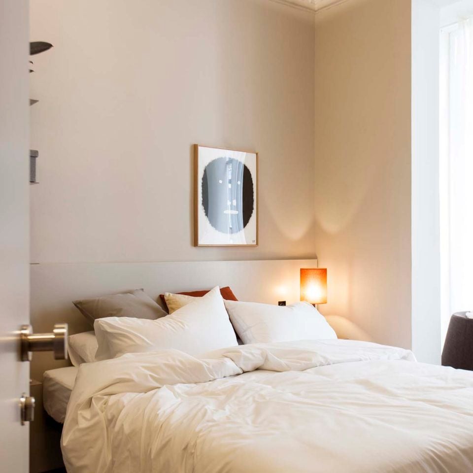 Modern Barcelona hotel room with minimalist design, white linens, wooden floor, large window offering city skyline view, sleek contemporary furnishings in soft neutral tones