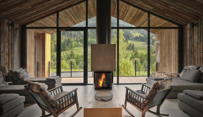 Modern luxury mountain resort with glass-walled infinity pool overlooking snow-covered Alps, surrounded by pine forest at sunset
