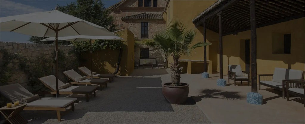 Rustic Spanish luxury hotel nestled in rolling countryside, featuring stone walls, terracotta roof, and elegant outdoor seating with mountain backdrop