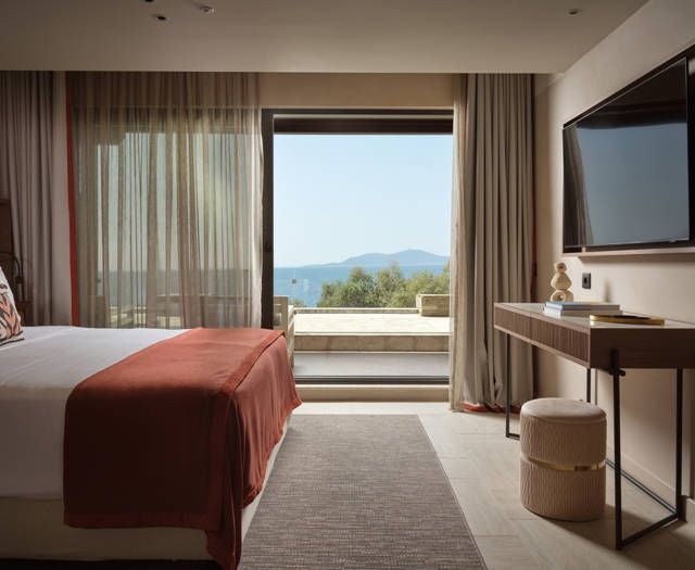 Luxurious junior suite overlooking azure Aegean Sea, featuring elegant white furnishings, private balcony, and panoramic coastal views of scenic Greece