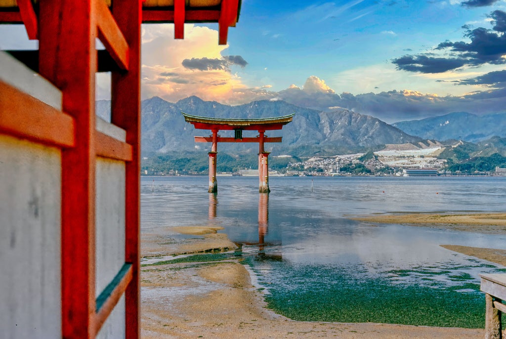 Iconic vermillion Torii gate standing in water at sunset, with Mount Misen and peaceful Miyajima island temple in background