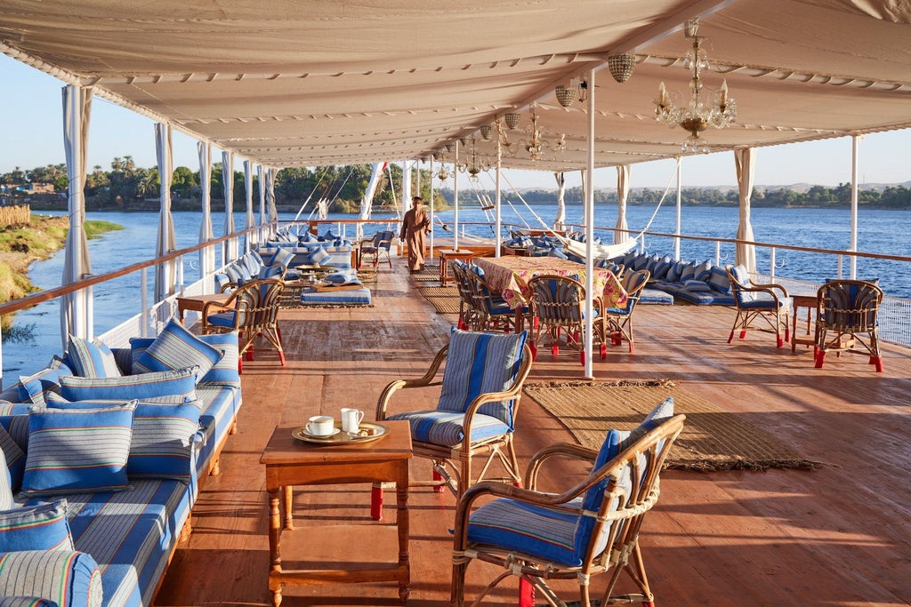 Traditional Egyptian wooden dahabiya boat with cream-colored canopy sailing on the Nile, featuring elegant deck furniture and panoramic river views