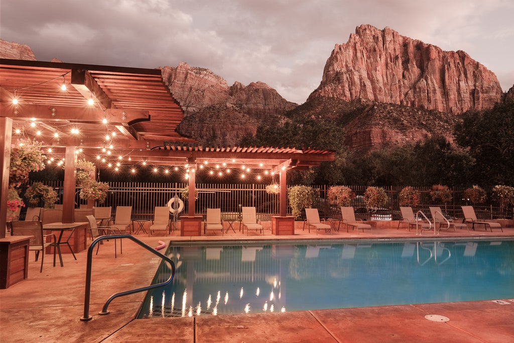 Modern mountain lodge with stone facade and glass balconies set against dramatic red cliffs, surrounded by lush landscaping at sunset