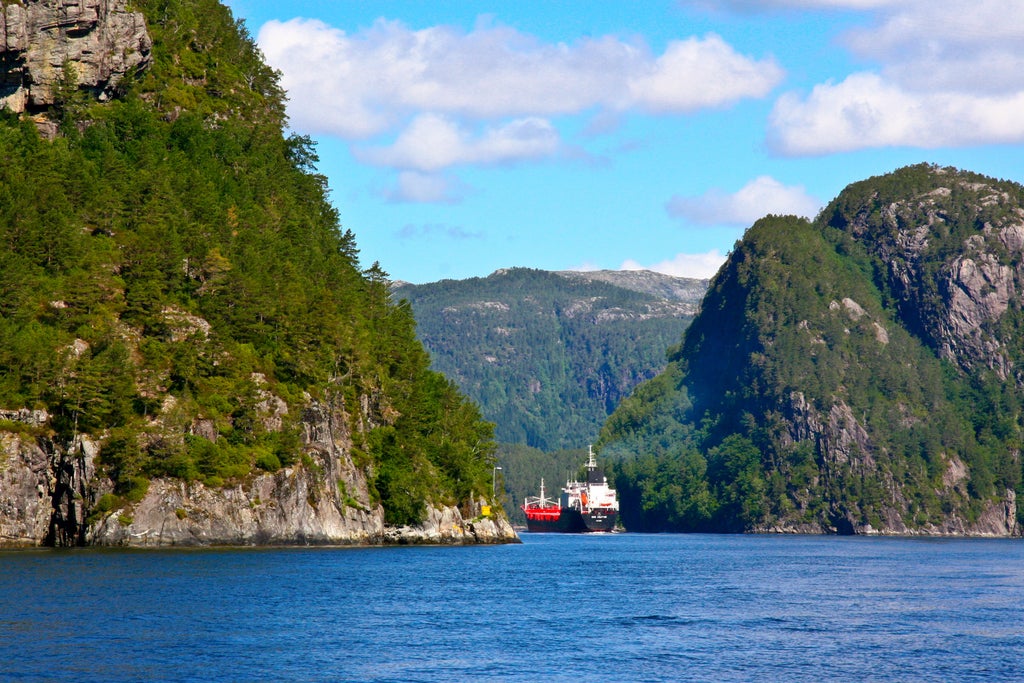 Small luxury boat glides through narrow Norwegian fjord, steep cliffs rise dramatically on both sides, calm waters reflect scenery