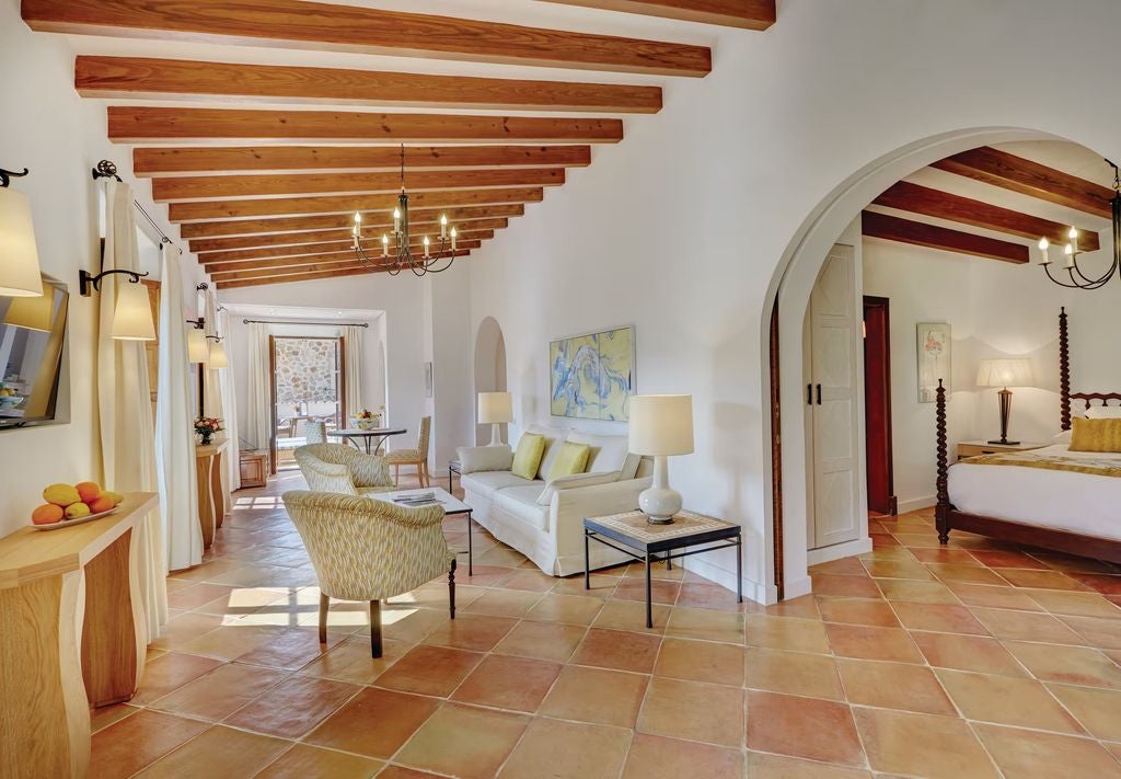 Elegant deluxe suite with terracotta floors, rustic wood beams, and a private balcony overlooking Mediterranean gardens in Mallorca