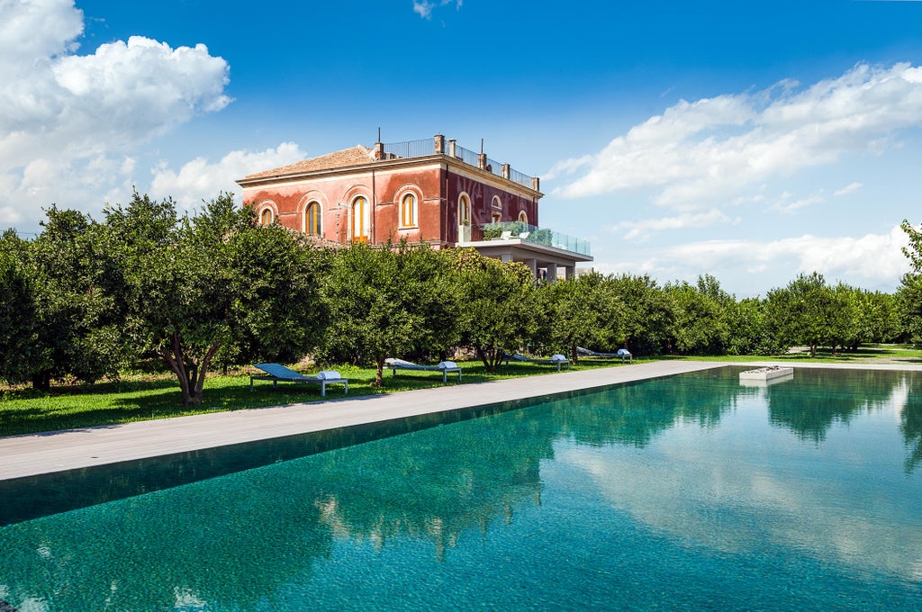 Elegant stone villa with infinity pool overlooking Mount Etna, featuring manicured gardens and Mediterranean architecture at sunset