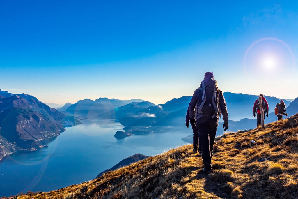 Snow-capped mountains reflect in Lake Como's crystal waters as hikers traverse scenic clifftop trails lined with Mediterranean foliage