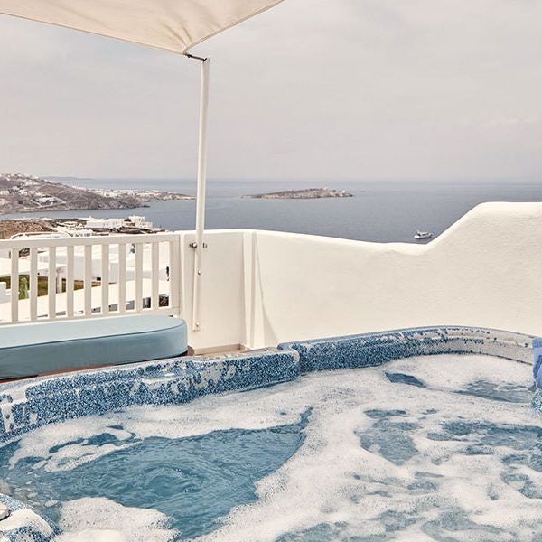 Luxurious white-washed sunset suite overlooking the Aegean Sea, featuring minimalist design with blue accents and panoramic ocean views from a private balcony in Mykonos, Greece.