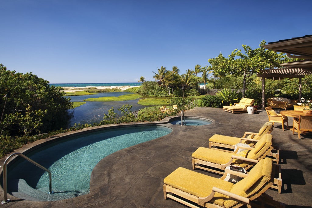 Oceanfront infinity pool surrounded by palm trees and lounge chairs at luxurious Hawaiian resort, with volcanic coastline at sunset