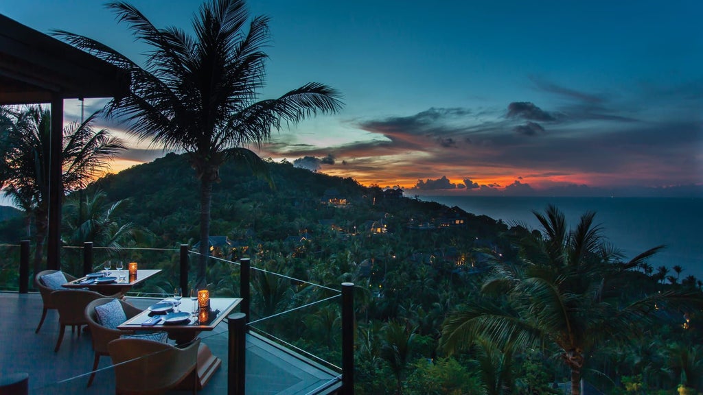 Luxurious infinity pool overlooking Koh Samui's turquoise ocean, with tropical palm trees and rustic wooden sun loungers at sunset