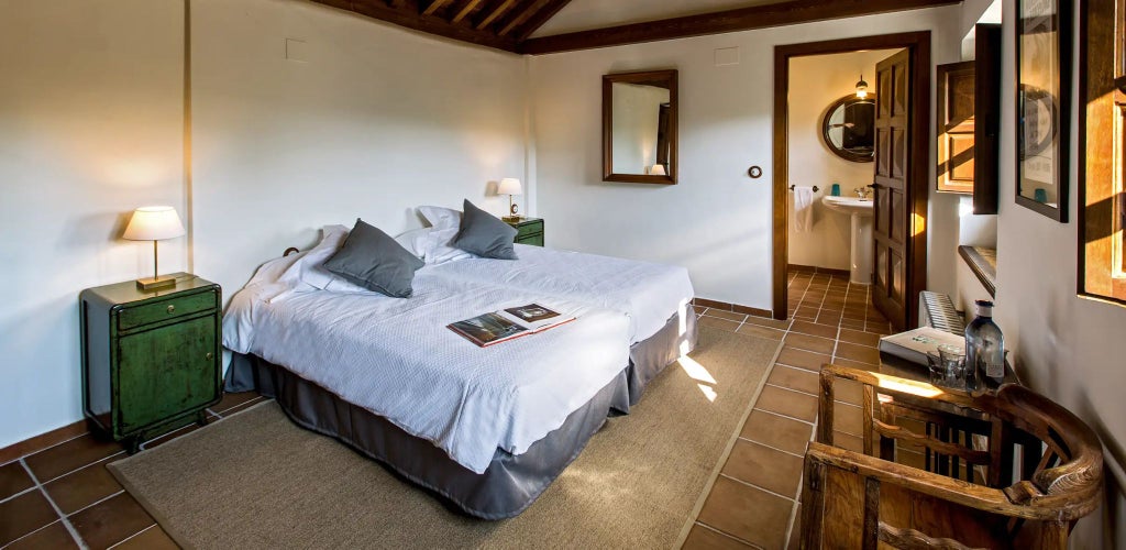 Elegant Spanish countryside bedroom with rustic wooden furnishings, soft white linens, and large windows overlooking rolling hills at a luxurious rural estate