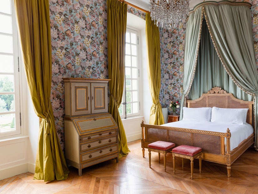 Elegant French château hotel room with ornate period furnishings, king bed, garden views through tall windows, antique artwork and crystal chandelier