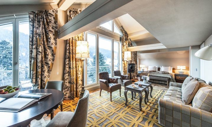 Luxurious Swiss hotel suite with panoramic mountain views, elegant white decor, king-sized bed, and floor-to-ceiling windows overlooking alpine landscape