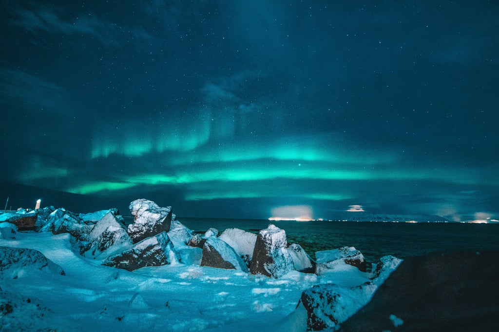 Northern Lights dance across dark Norwegian sky above luxury cruise ship, its warm lights reflecting on calm fjord waters below