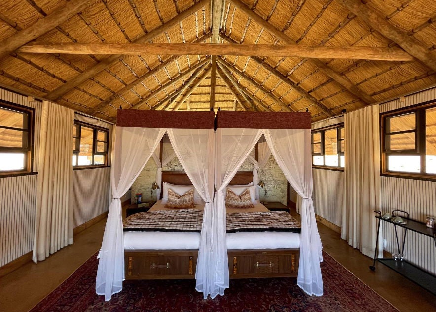 Two luxurious single beds with crisp white linens in desert lodge room, featuring canvas walls and expansive mesh windows with dune views