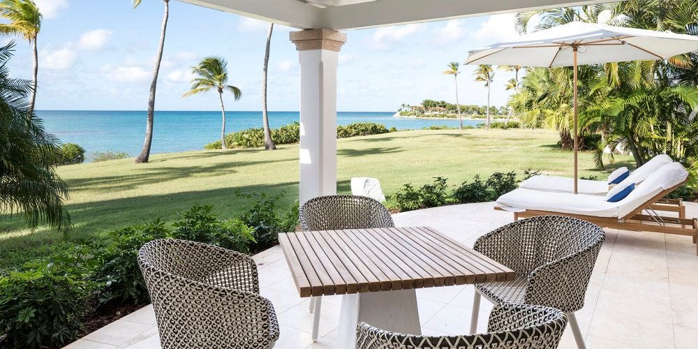 Luxurious beachside suite at Jumby Bay with pristine white linens, elegant four-poster bed, and panoramic ocean view through floor-to-ceiling windows