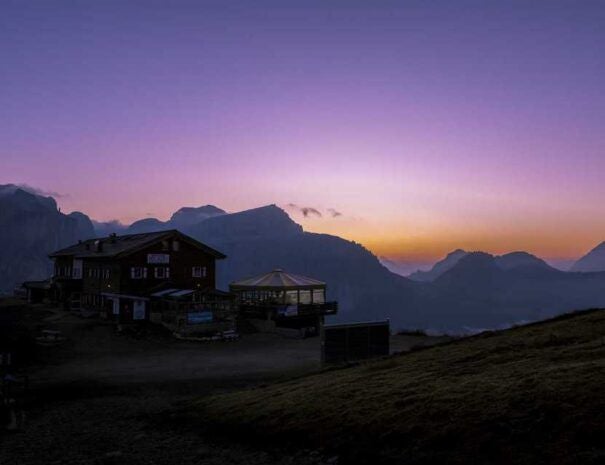 Stone and timber Alpine lodge with snow-capped mountain backdrop, rustic wooden balconies and glowing warm windows at dusk