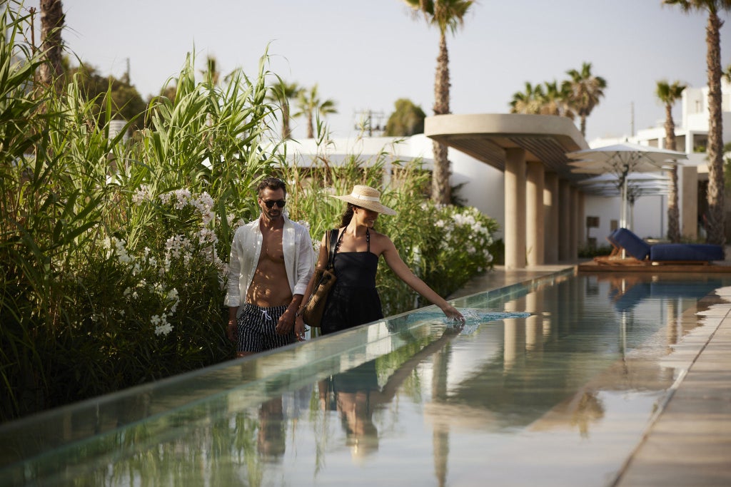 Whitewashed beachfront hotel with infinity pool overlooking turquoise Aegean Sea, surrounded by Mediterranean gardens and private cabanas