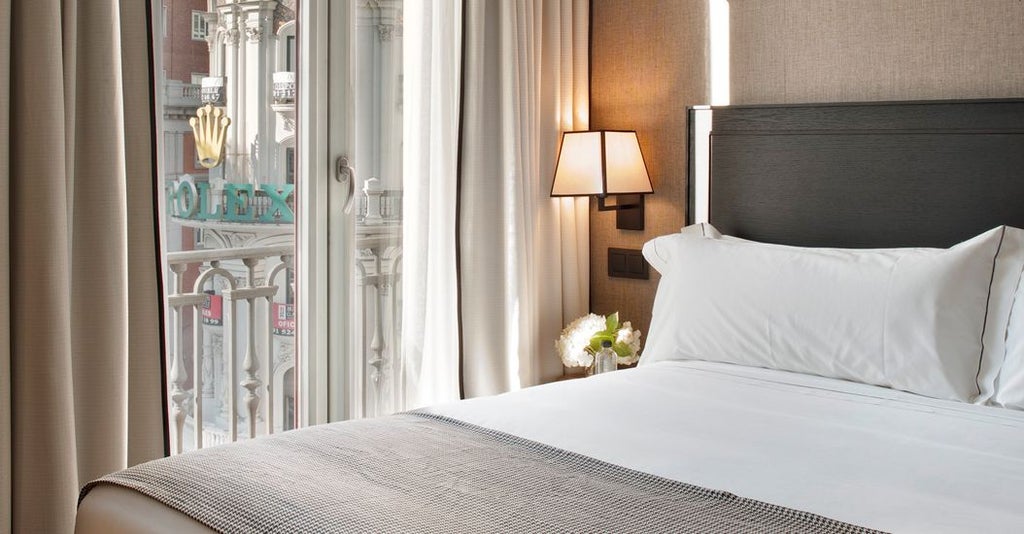 Luxurious deluxe hotel room with floor-to-ceiling windows, elegant modern decor, and panoramic city views of Madrid's stunning urban landscape at sunset