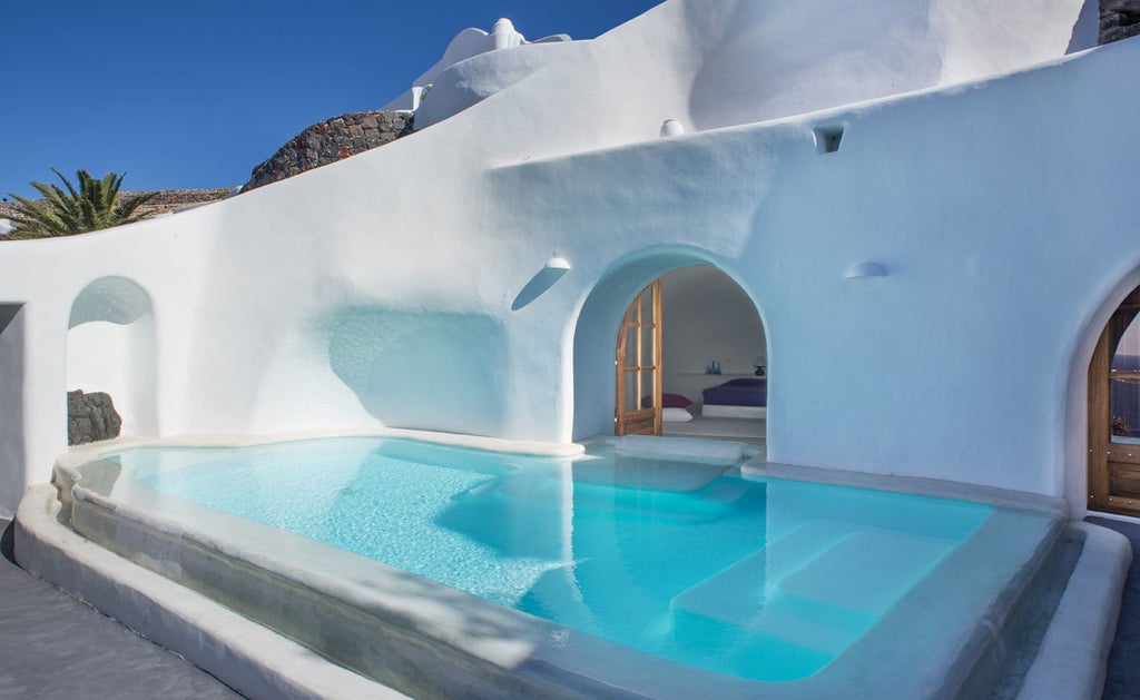 White-washed luxury cave hotel built into Santorini cliffs, featuring curved archways and infinity pool overlooking Aegean Sea at sunset